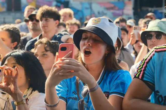 A woman wearing a football shirt and summer hat sings from the crowd as she records on her phone.