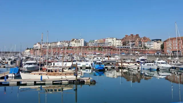 Several boats ranging in different colours and sizes are moored in a harbour in Kent. The sea is still and the sky can be seen reflected in the water, with a long row of buildings seen in the background