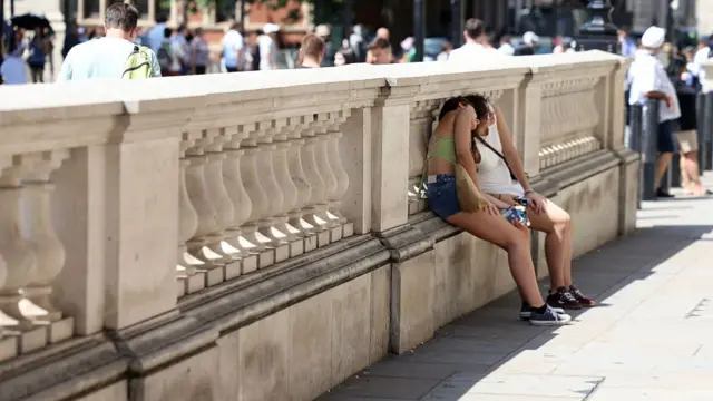 Two people sit on a step, hiding underneath a banister above it to remain in shade while the sun beats down above them.