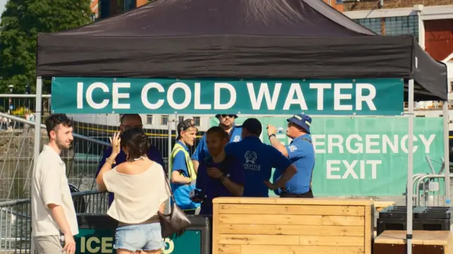 Two concert-goers stand in front of a gazebo with the words 'ice cold water' on it. A group of staff can be seen behind the stand.