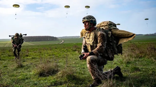 Two soldiers on the ground in full gear on Salisbury plain. Parachutists can be seen descending in the background.