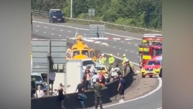Emergency services and motorists on motorway. There is a yellow helicopter nearby and two fire brigade vehicles.