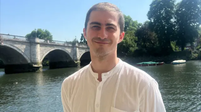A man wearing a white top, smiling, while standing on a river boat by the river.