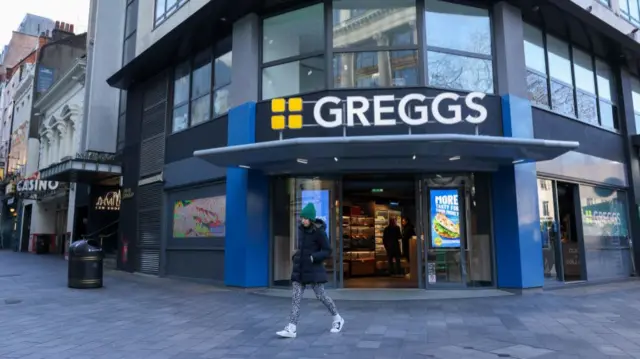A woman wearing a padded black coat, spotty patterned tights and a green beanie hat walks past a Greggs store on the corner of Leicester Square