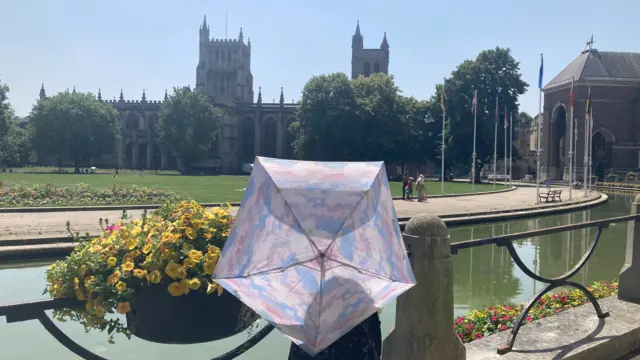 A person looks over Bristol's College Green and cathedral with a raised purple umbrella obscuring the back of their head. It is very sunny, with a clear blue sky.