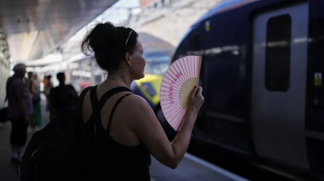 A woman standing on a train platform using a fan to cool herself.