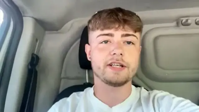 A man wearing a light coloured top, sitting in a vehicle.