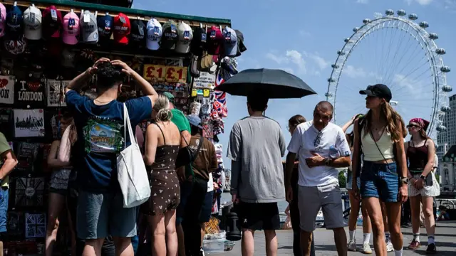 People in London stand beside a souvenir stand in London. The London Eye is visible in the background.