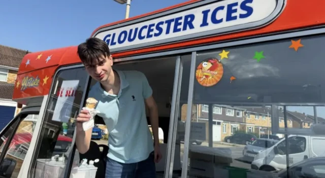 A teenage boy leans out of the window of an ice cream van, he is holding an ice cream in a cone.