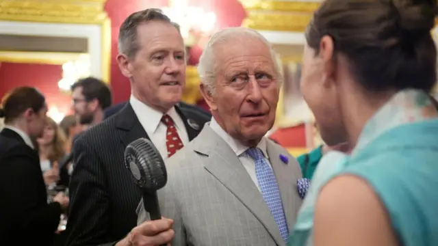 King Charles III rolling his eyes in a room crowded with people as a man stands behind him holding a small fan close to his face