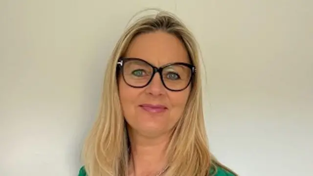 A headshot of a woman with straight, long blonde hair. She is wearing spectacles with a dark, thick rim and is smiling.