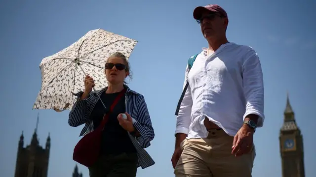 A woman and a man walk in central London on a sunny day. The woman is holding an umbrella to shield themselves from the sun.