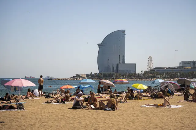 People sat on a beach, shading from the sun with umbrellas and sat on towels. The sea and some large buildings are in the background.