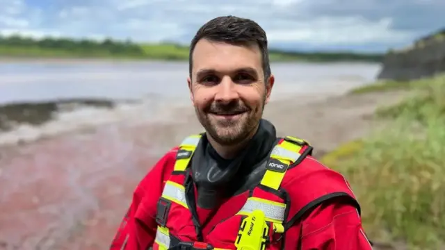 Matt smiles at the camera wearing his red and yellow rescue uniform. A river beach can be seen behind him.