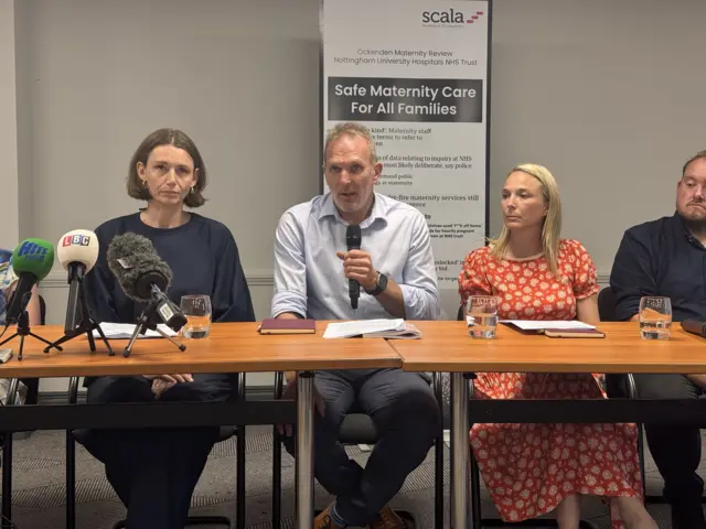 Two men and two women sat behind a long desk, one man is holding a microphone, looking at the camera.