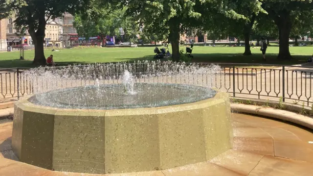 A large stone fountain sprays water into the air. Behind it there are people resting under the shade of the trees.