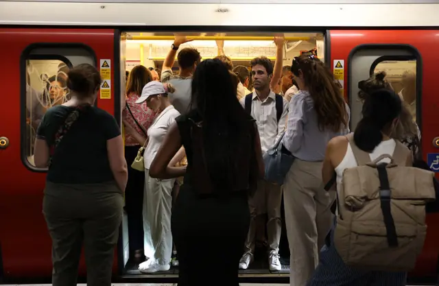 Lots of commuters packed into a tube carriage, with red doors open and people holding onto the yellow bars.