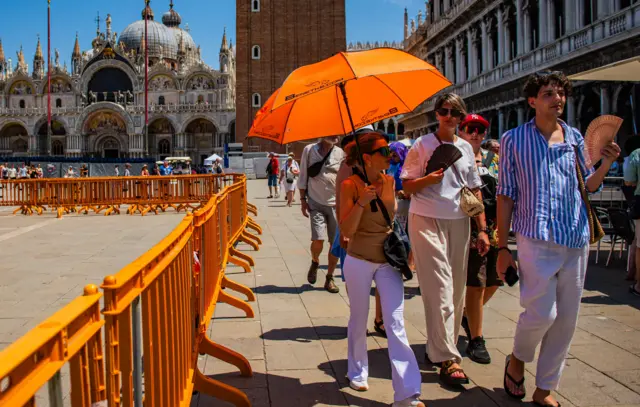 Tourists holding an orange umbrella, wearing sunglasses and having handheld fans. There are orange gates around the square and large buildings in the background.