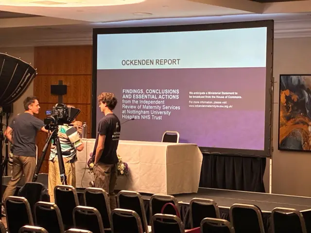 A camera is set up in front of a table on a raised stage. A presentation screen reading "Ockenden report" is behind.