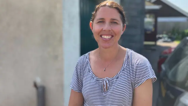 Hayley has brown hair that is tied back in a ponytail. She is smiling at the camera and wearing a striped top with a necklace.