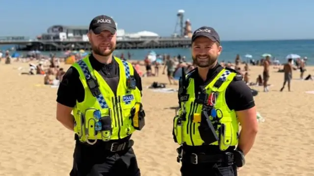 Two officers on a busy beach, a pier in the background.
