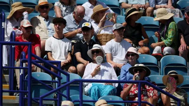 A crowd is sat in plastic seats watching a tennis match as the sun bears down on them. There is a mix of ages from old to young, with many people wearing hats and sunglasses, while one woman holds an electric fan to her face