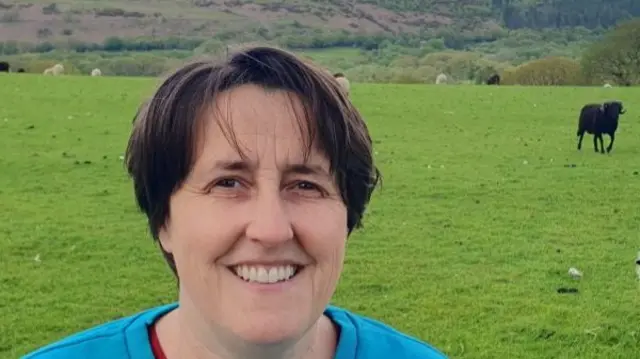 A woman in a blue sweatshirt and red t-shirt underneath smiles at the camera, she has short brown hair. In the background is a grass field with a black sheep