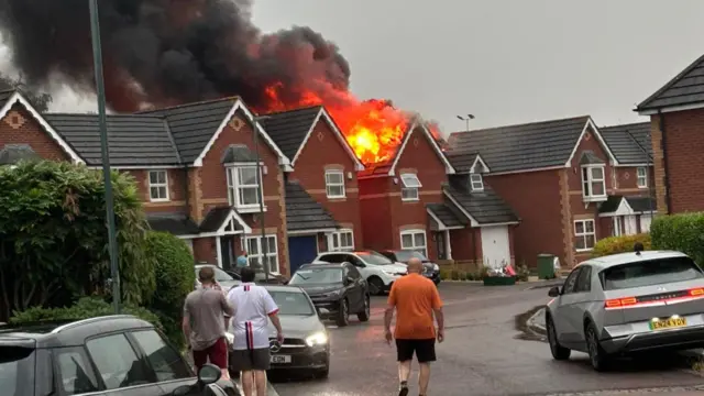 A detached house fully alight on a residential street, as neighbours look on.