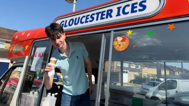 A man wearing a light blue top, holding an ice cream, while in an ice cream van.