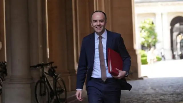 James Murray walks out onto Downing Street, he wears a blue suit and brown tie and holds a red folder under his arm