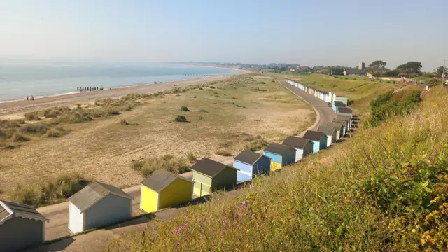 A line of different coloured beach huts curve along a concrete road very close to the shoreline. In front of them is a dry grass field, which is dotted with a few bushes leading towards the beach and the sea in the distance