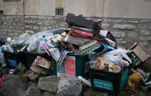 Recycling bins piled high with waste sit on the pavement.