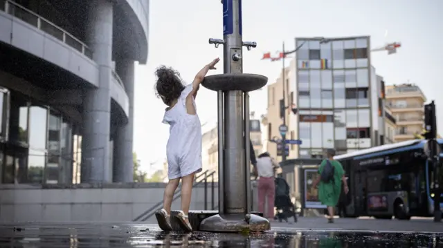A child, wearing white, reaches for the water coming out of a silver water fountain, with puddles of water around the fountain.