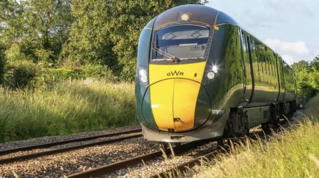 A GWR train, which is yelow and dark green, travels along a rural track surrounded by grass and trees. It is a sunny day.