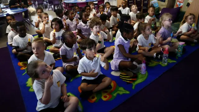 A classroom full of young children sitting cross-legged with ice lollies in their hands and mouths. They are all in school uniforms.