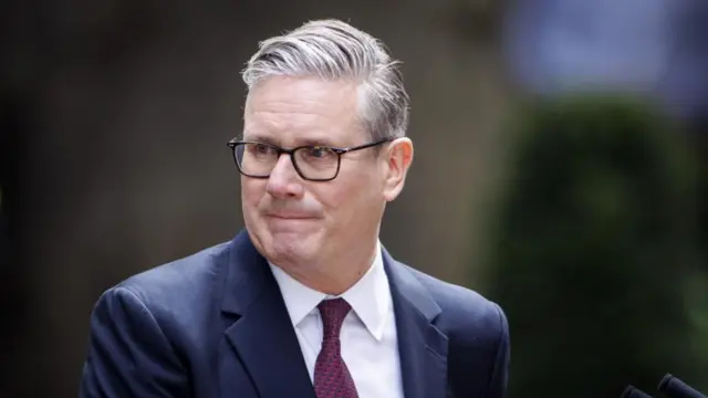 Keir Starmer has grey hair and wears glasses, a white shirt, burgundy tie and a blue jacket