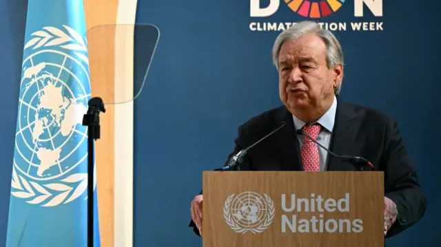 A man with grey hair wearing a black suit with a red tie stands at a podium that reads "United Nations" behind his right is a UN flag and behind his head a sign that reads London Climate Action Week