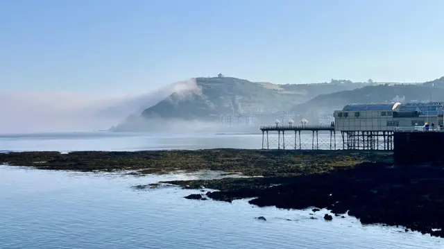 A pier is stood on a headland jutting out to sea in Wales. In the distance a large cliff rises above several houses along the coastline, while a layer of fog extends from the right
