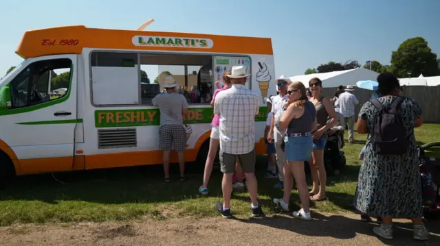 People queue in line at an ice cream van, which is parked on a field next to a dirt path in the sun. A couple of white gazebos can bee seen in the background to the right below a clear blue sky