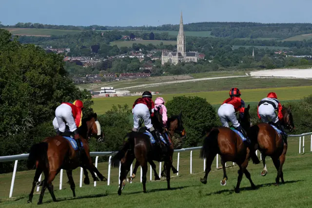 A group of horses and riders head towards a bend at Salisbury racecourse on a sunny day. Salisbury Cathedral is visible in the distance