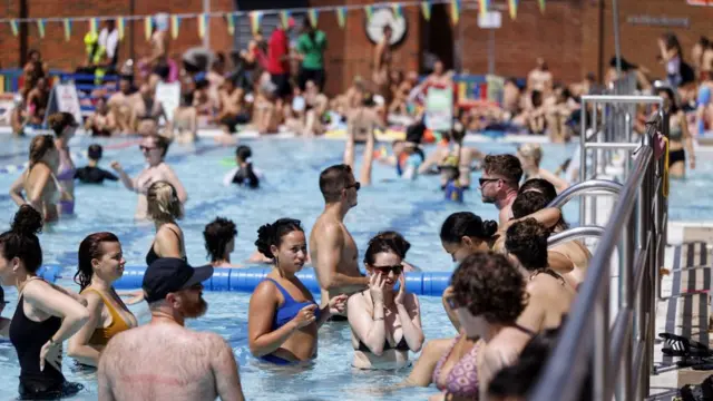 Multiple people can be seen in a packed outdoor lido. A line of people can be seen sitting on the edge of the pool in the blurred background.