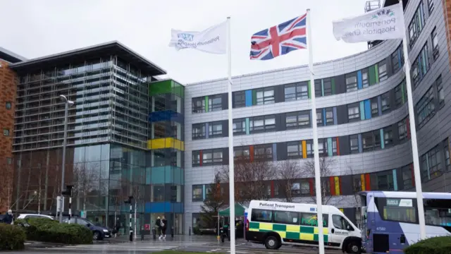 A view of the Queen Alexandra Hospital, with an ambulance transport with yellow and green square patterns, and three flags at the top, including the union jack.