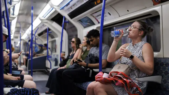 A woman wearing a black and white spotted dress sips from a plastic water bottle while sat down on the tube. She is surrounded by other passengers some of whom are on their phones or holding coffee cups