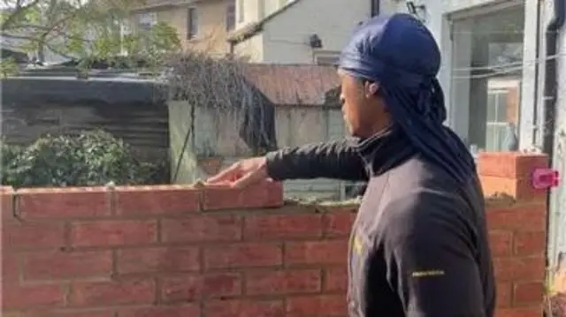A bricklayer places a brick on the top of a brickwall being constructed. He is wearing a dark zip-up jumper and the day appears to be sunny.