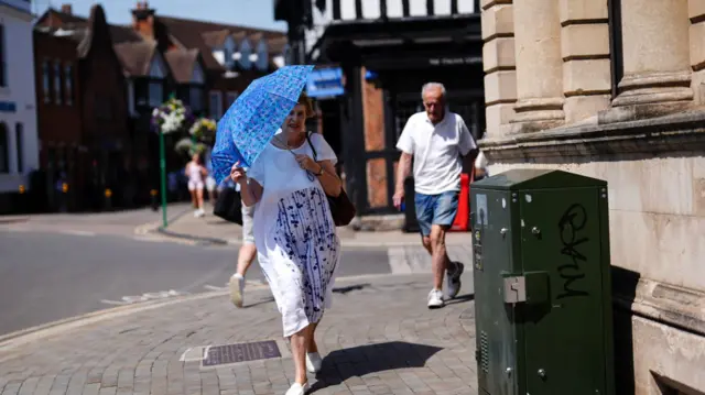 A woman shields herself from the sun using a blue umbrella as she walks through a high street.