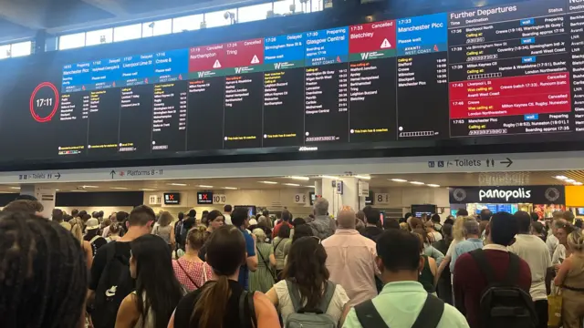 A departure board at London Euston, being looked at by lots of commuters.