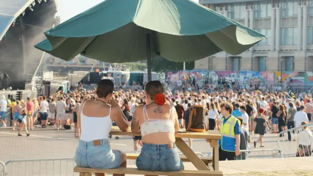 Two women wearing vest tops and shorts sit under a parasol on a bench looking out over the gathering crowd.