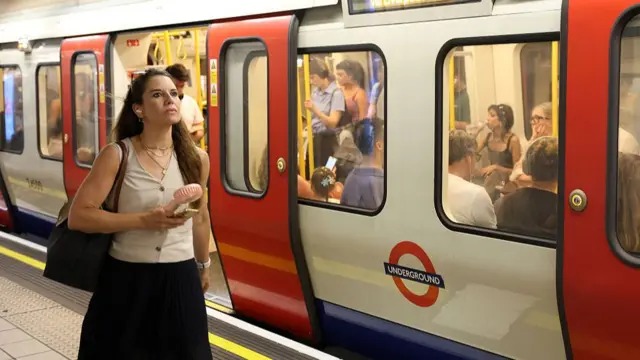 A woman wearing a white sleeveless top and black dress blow a portable fan in her faces while walking along a tube platform. A Tube train full of commuters is stationary on the platform next to her