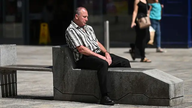 A man in a striped shirt and dark trousers sits on a concrete bench on a sunny day.