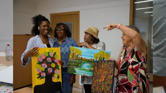 Four women stand in a row smiling, they are holding up paintings they have made. On the left is a younger woman with brown hair wearing a blue and white shirt and white headphones in. To her left are three older women, the first is in a blue shirt and blue jeans with glasses, the second is in a white shirt with a brown sunhat and the third is in a multicoloured floral dres